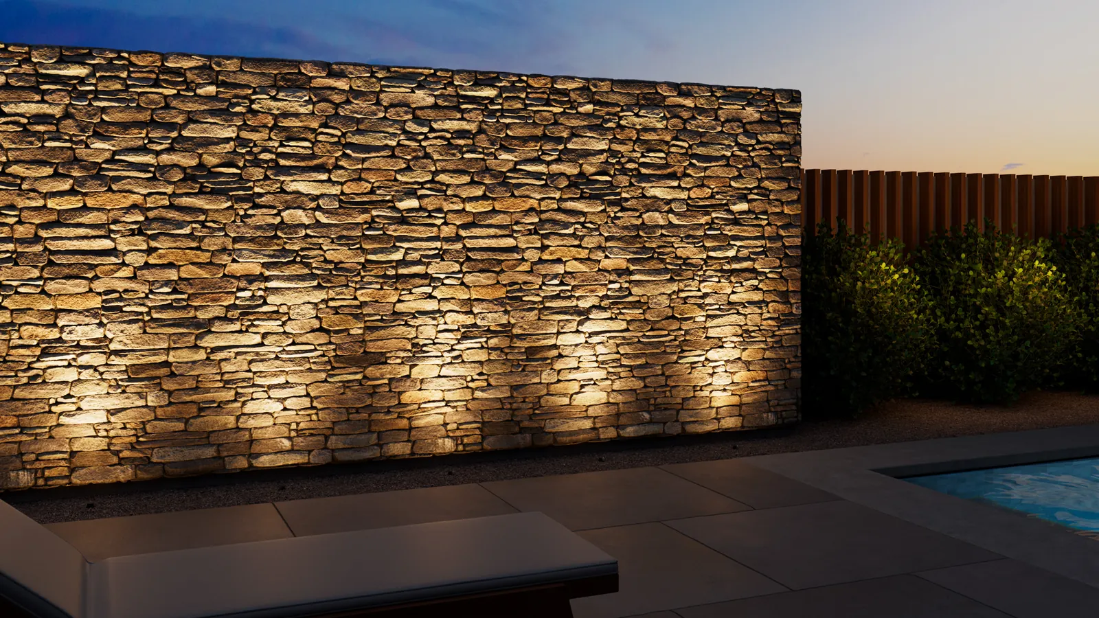 Stacked stone wall grazed with warm light from in-grade fixtures beside a pool at dusk